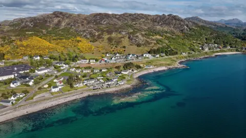 An aerial shot showing blue water in front of a village with a green and brown hill in the background. Yellow gorse bushes fill the left hand side of the hillside, with several houses and buildings in front of a rocky shoreline. 