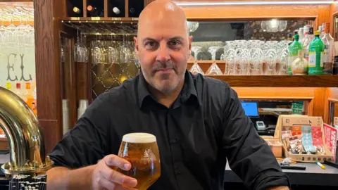 Darren Summers holds up a pint of beer while standing behind a pub bar
