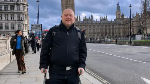 Martyn O'Brien, un hombre calvo de mediana edad que se apoya en muletas y posa para una foto frente al Palacio de Westminster.