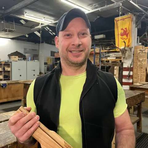 Designer Tim Norman holding the wooden baton. He is standing inside his workshop and is wearing a blue cap, black jacket and yellow T-shirt