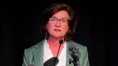 Eluned Morgan stands against a black back drop and speaks into a microphone. She wears glasses, a white shirt, and green blazer. She has shoulder-length brown hair. 