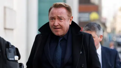 Michael Flatley. He has short reddish hair, wearing a navy suit and black coat.