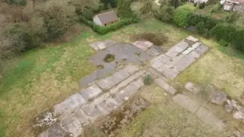 An aerial view of a piece of grassland with the stone remains of the foundation of a building. There are other buildings beyond a hedge nearby.