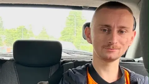 Rachael Williams Daniel is sat in the back of a car looking at the camera. He has a shaved head and Wolves top on under a jacket.