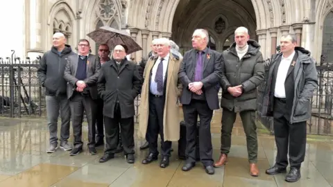 A group of men in smart suits and coats stand outside the entrance of the High Court, a grand, gothic building. They look solemn, and it appears to be raining.