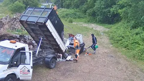 Two people are standing around a van which is disposing of various waste such as an oven cooker and a sofa. The van is on grassy land.