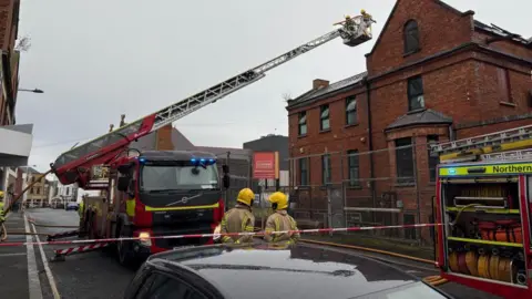 A fire appliance being used to fight the fire in the affected building. Fire officers are standing on the ground wearing yellow helmets. 