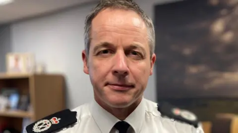 Paul has short receding grey hair and hazel-blue eyes. He is wearing a police uniform consisting of a white shirt with badges on the lapel and a black tie