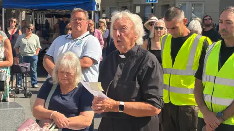 BBC/Ewan Murrie Reverend Pat Alden during the community vigil in Shoreham-by-Sea on 22 August. She is dressed in typical vicar attire and is holding a piece of paper.