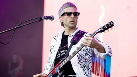 An image of the Irwin Sparkes, lead singer of the Hoosiers, playing a guitar wearing sunglasses and a jacket with fringing on the sleeves. 