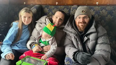 Lucy and Josh with their son Freddie and his sister are sitting on a railway seat. Freddie is wearing an elf hat and looking towards the window.