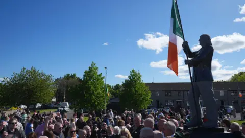 A crowd gathered around a statue of Bobby Sands, with an Irish flag hanging on it. The sky is clear and blue above. There are some trees and a row of houses in the background. 