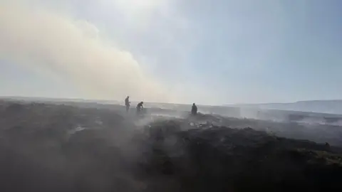 Glossop Fire Station Three firefighters can be seen in the distance damping down a moorland wildfire. Plumes of smoke can be seen rising from the ground