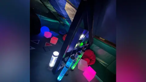 New Reflexions The corner of a room is illuminated by various lava lamps and a glowing cubes in assorted colours such as red, pink, white, blue and green. There is a dividing partial, made of individual sections of wood. The ceiling is sloped, as if the room is in an attic, with blinds drawn on what look like Velux windows. Because of the sloped ceiling the far wall isn't very high and is covered in green padding.