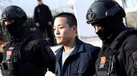 Getty Images A man being led away by two armed guards wearing heavy duty armour, bulletproof vests, helmets and balaclavas.