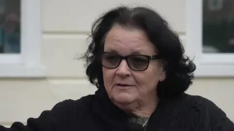 BBC/Chrissie Reidy A dark-haired woman wearing dark glasses and a black jumper.