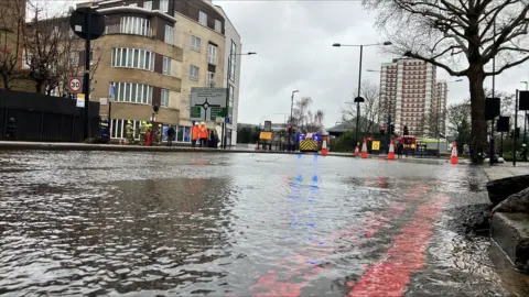 A road covered in flood water with cones and a fire appliance in the background. 