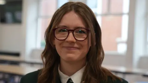 A teenage girl with long brown hair, wearing glasses, a white shirt and a green blazer. She is standing in a canteen, with tables, a white wall and large windows behind her.