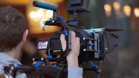 A close-up view of a professional video camera being operated indoors. In the foreground, a person is seen from behind, holding and operating a large camera rig. The focus of the image is the camera equipment rather than the person.