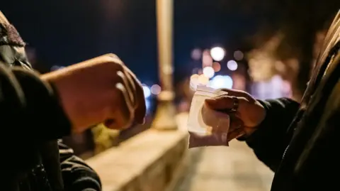 Close up shot of a bag of a powder in a small clear bag being passed to a man on the street. It appears to be dark and there are some lights in the distance.