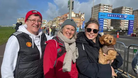 Helen Andrews, Tracy Gosney and Grace Harman wearing jackets by the side of a road, smiling at the camera. Grace Harman, on the right, is carrying a small brown dog. A sign reading Brighton & Hove 10K is visible in the background.