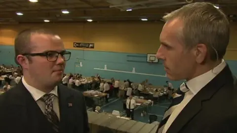 Two men talking on camera. The man on the right is interviewing the younger man on the left.