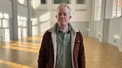 Man with short grey hair in denim shirt and sheepskin jacket stood in hall