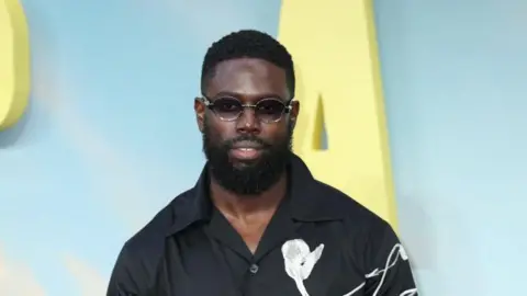 Getty Images Ghetts wears sunglasses and a black shirt with a white flower motif