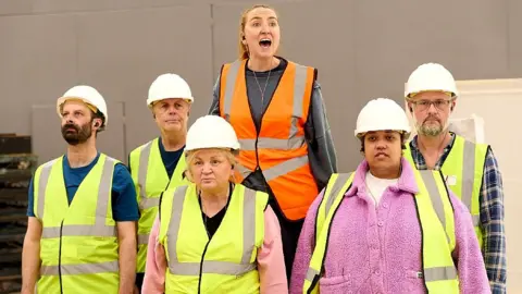 Mark Douet Six actors stand in a group. Five wear yellow high-vis jackets and white hard safety helmets. A sixth is standing in the back on something making her higher than the others. She wears an orange high-vis jacket and no helmet.