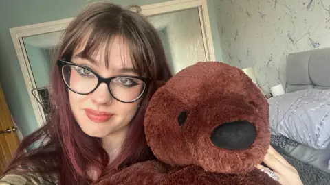 PA Media Cara O'Brien is on the left of the image, holding the teddy bear next to her head. She has red hair, and is wearing black glasses smiling at the camera. On the right, next to her, is the brown bear that has black eyes and nose. 