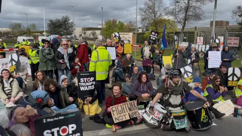 A group of about 50 people sit on the road holding signs with messages including Stop Trump's Wars, and No War on Iran. To their right is the base's wire fence.