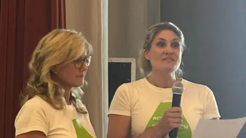 Two women wearing white t-shirts with green logos on them are standing. One of the women is holding a microphone and a piece of paper.