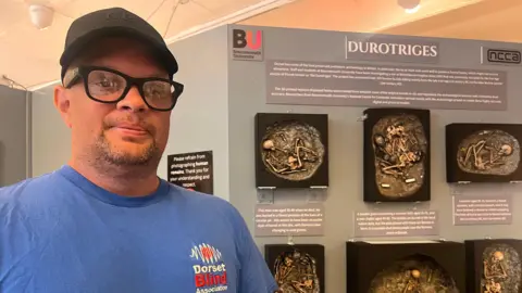 Darrel Bennett poses for the camera alongside mounted 3D exhibits. He is wearing a black cap, black-rimmed glasses and a blue t-shirt with a Dorset Blind Association.