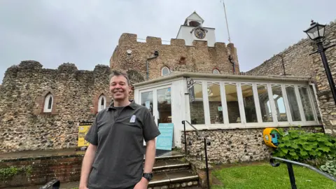 Lewis Macleod is standing outside the Clock Tower cafe in a black polo shirt.