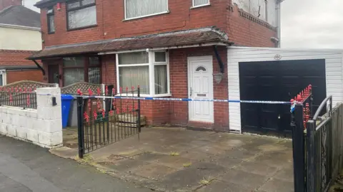 Blue and white police tape is attached to gate posts of a driveway in front of a house. There is a garage with a black door and a white front door to the property, which has a bay window.