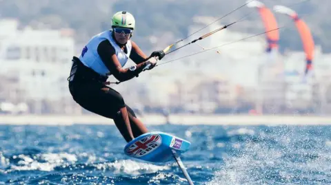 Lily Young is pictured kiteboarding. She is stood on a board on a body of water and gripping a beam that is connected to several wires. She board is airborne and a fin can be seen on its underside. She is facing in the direction of the camera and is wearing a helmet, goggles and a life jacket. 