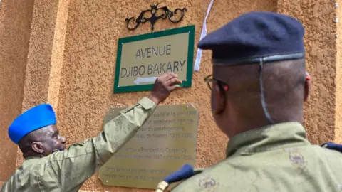 AFP Um oficial militar de uniforme verde e boné azul, inaugurando a nova placa com os dizeres Avenida Djibo Bakary