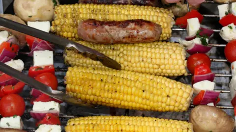 Barnaby Perkins/BBC A close-up of a barbecue grill with a line up of food resting on the grill. There are skewers with tomatoes, cheese, onions and potatoes and a sausage resting on one of four sweetcorn cobs. Tongs are reaching in to move a sweetcorn cob. 