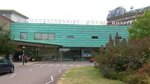 A picture of the exterior of Gloucestershire Royal Hospital. The front of the building ins clad with green panelling and large letters spell out the name of the hospital along the roof. 