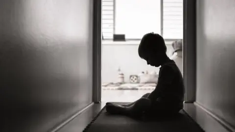 A young child is sitting alone in a hallway in front of an open door.