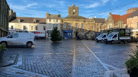 Alnwick's cobbled market square surrounded by Georgian buildings. In the middle stands a small, undecorated Christmas tree dwarfed by its surroundings.