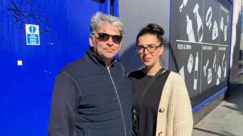 Alex Pope/BBC A man and a woman, standing outside a B&M store. The man has on a blue coat, with sunglasses on and grey hair. He is standing next to a woman with a black top on, cream cardigan, hair up in a bun, with glasses on and a bag across her. They are by a blue door, with graffiti on it and another sign.