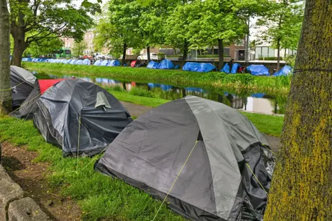 Getty Images Two rows of tents - 1  grey, 1  bluish  - are pitched on  some  sides of a canal, nether  trees. 