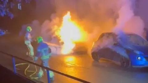 Blue car on fire, with firefighters with a hose trying to put the blaze out.