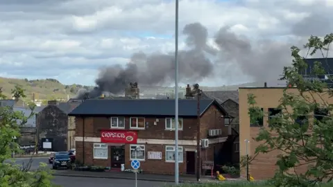 A street scene with a brick commercial building in the foreground displaying a red sign. Thick, dark smoke rises from behind the buildings, drifting across the sky. Parked cars, a lamppost, and traffic signs line the road, with hills and scattered buildings visible in the distance under a partly cloudy sky.
