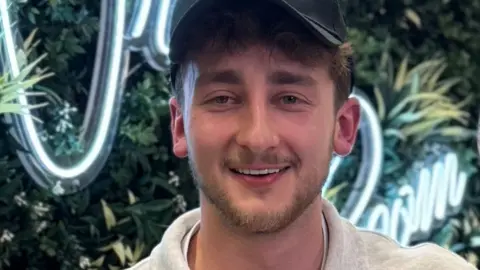 Brandon B smiles wearing a black cap while standing in front of a green leafy wall
