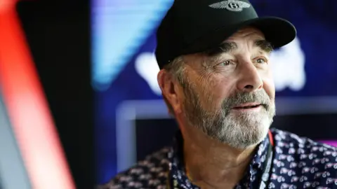 Nigel Mansell is wearing a black cap and a patterned navy shirt, and looks off to the side of the camera. 