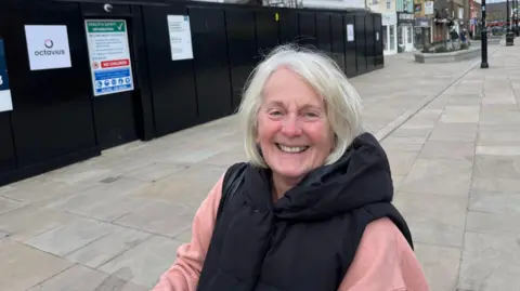 John Devine/BBC Kate Isaacs has light blonde shoulder length hair, she has a black body warmer with a pink jumper underneath, behind her is 2m (6 ft) tall black security fencing around a demolition site, with part of an old building still standing.
