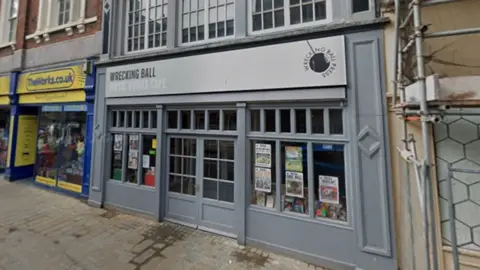 Google maps The front of the building is painted grey and has posters on the wall. The Works is next door and the shop at the other side has scaffolding in front of it.