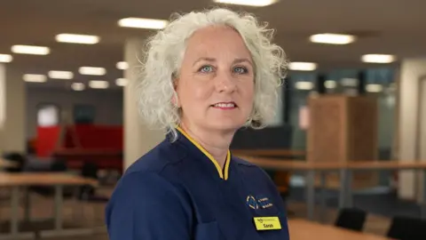 Sarah Fox, head of programme for Antenatal Screening Wales. She has white hair and blue eyes and is wearing a dark blue scrub top with yellow trim, with an NHS Wales logo 